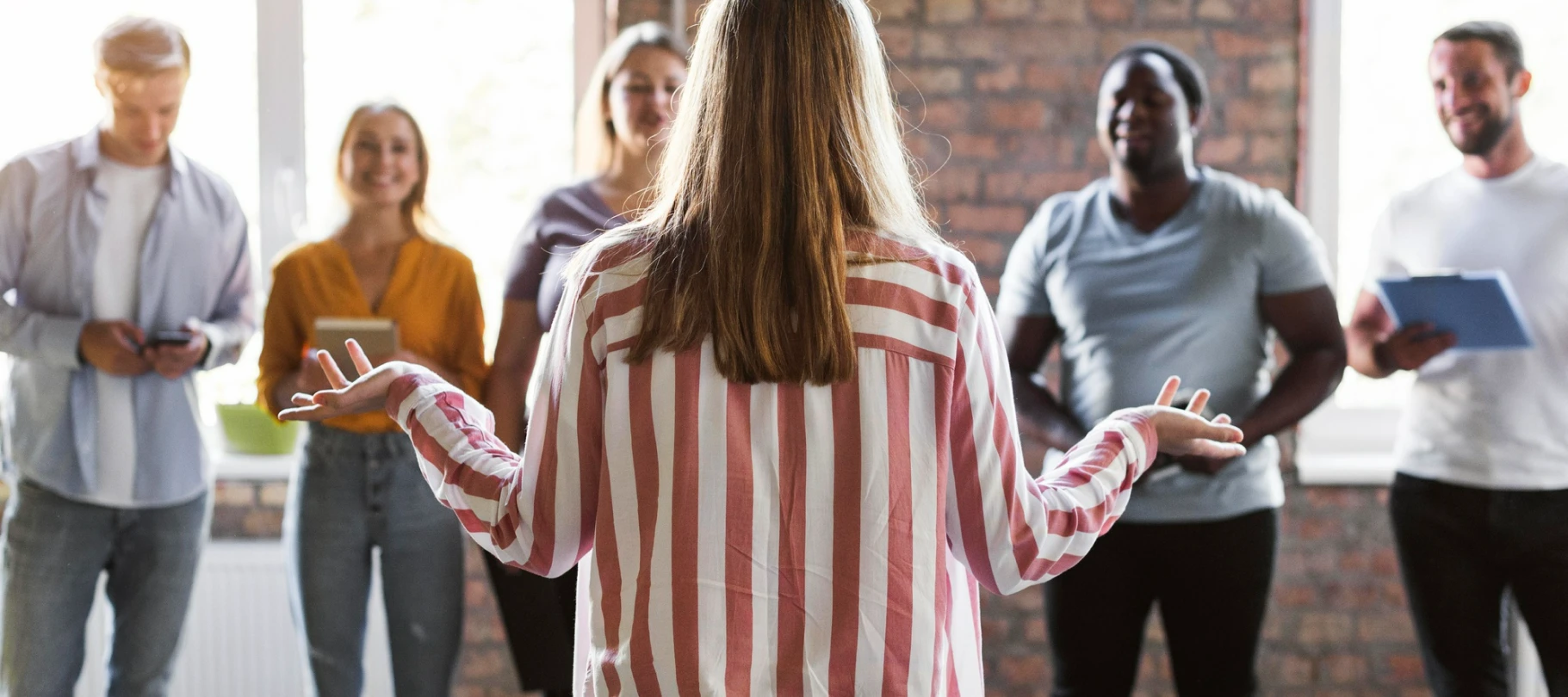 Mujer de espaldas dando una charla motivacional a un equipo diverso en una oficina moderna.